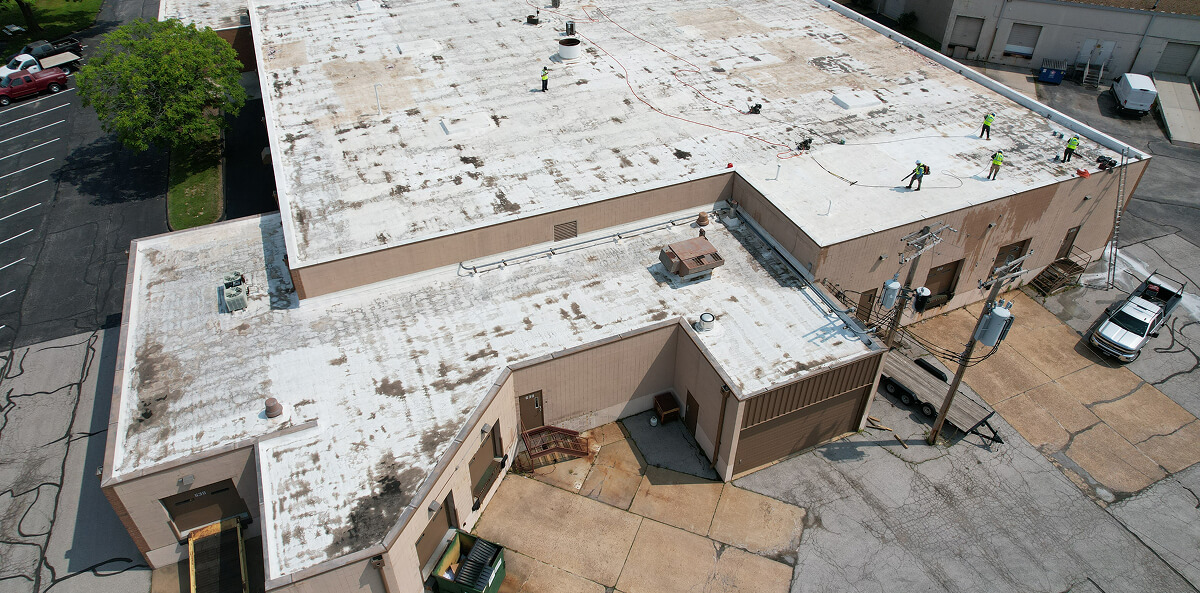 Aerial view of crew in safety gear performing TPO flat roof restoration on a large commercial building with a flat roof and empty parking lot.